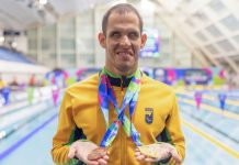 Vestindo a jaqueta amarela do Brasil, com a as piscinas ao fundo, Matheus Rheine exibe a medalha de bronze na mão direita e a de ouro na mão esquerda, no Mundial de Natação Paralímpica em Manchester, 2023