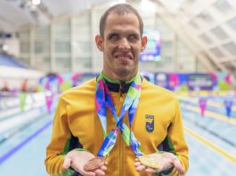 Vestindo a jaqueta amarela do Brasil, com a as piscinas ao fundo, Matheus Rheine exibe a medalha de bronze na mão direita e a de ouro na mão esquerda, no Mundial de Natação Paralímpica em Manchester, 2023